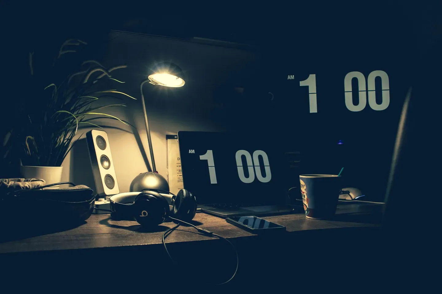 A dark home desk lit by a lamp, with a laptop and a screen both showing 1:00 AM